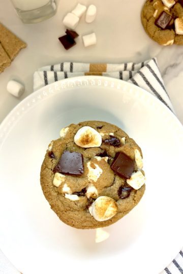Chocolate chip s'mores cookie on plate over dish towel next to glass of milk and marshmallows around