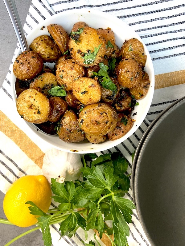 Oven roasted mini potatoes in a bowl on dish towel next to lemon garlic and parsley