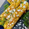 Four grilled corn on the cob with mayonnaise and feta cheese next to parsley served on board