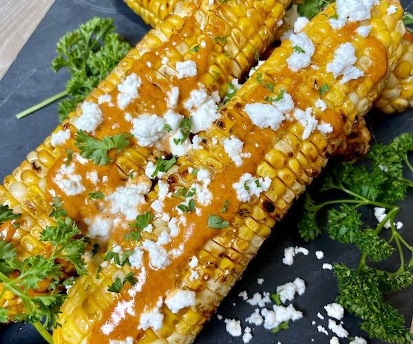 Four grilled corn on the cob with mayonnaise and feta cheese next to parsley served on board