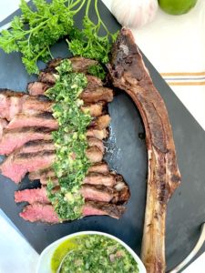 Presentation of sliced tomahawk steak on board with chimichurri sauce next to bone with a bowl of chimichurril