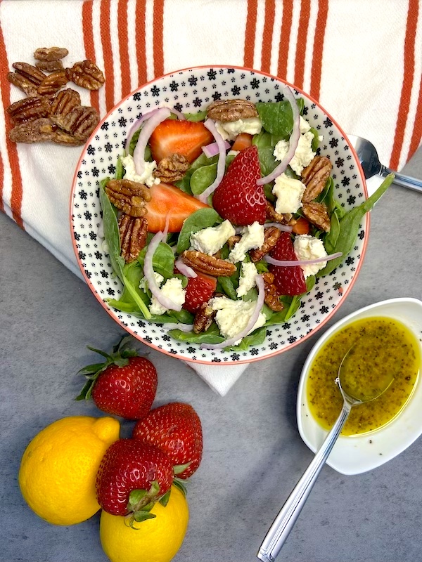 Presentation of salad bowl on dish towel next to maple pecans, strawberries, lemon and salad dressing