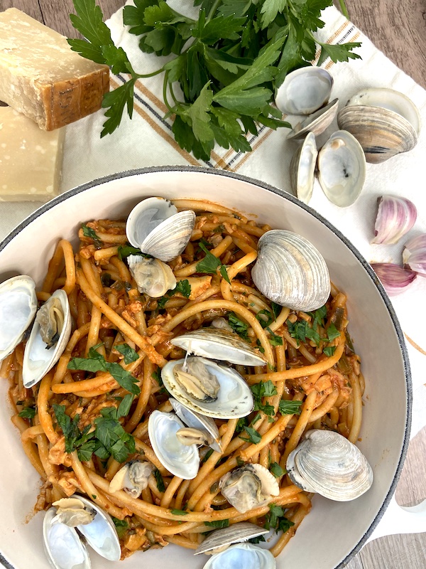 Presentation of prepared bucatini alle vongole with clam shells next to garlic cloves parsley and cheese block
