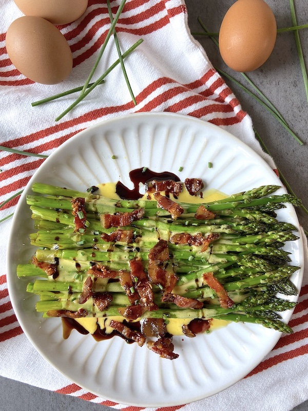 Plate of asparagus ready to be served, with hollandaise sauce, drizzle of balsamic vinegar and topped with guanciale.