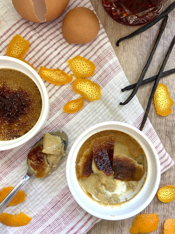 Image of two crème brûlées where one is eaten and th other one intact. They are next to cracked eggs, orange peels and 4 vanilla beans.